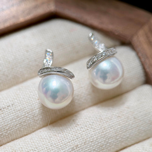 A pair of silver stud earrings with large white freshwater pearls and a curved row of small clear stones, displayed on a cream fabric surface inside a wooden box.