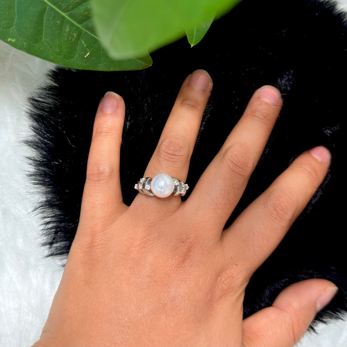A silver ring with a large white pearl and small clear stones on the ring finger of a hand, resting on a black furry surface with a white furry surface underneath and green leaves at the top.