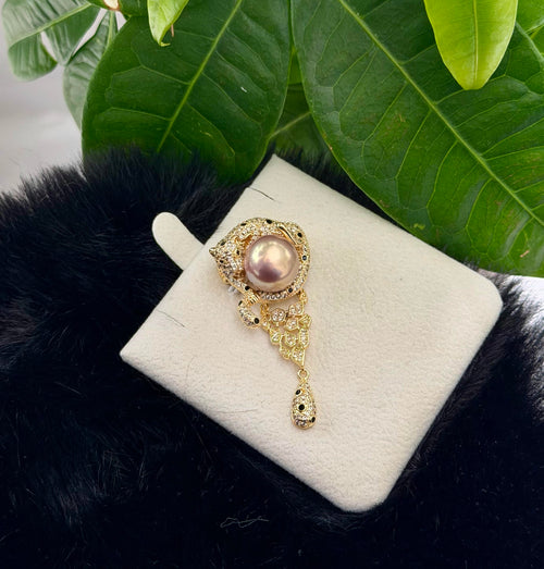 A gold-toned ornate brooch featuring a central pink pearl and small clear stones, displayed on a cream jewelry card over a black faux-fur surface with green leaves in the background.
