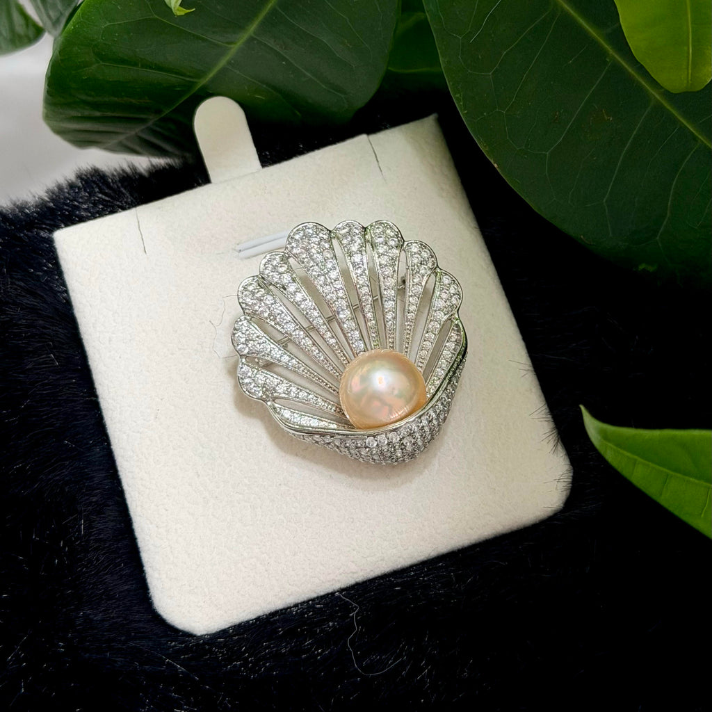 A shell-shaped silver brooch with a central pearl and pavé-set clear stones, displayed on a white jewelry card with green leaves and black fabric in the background.