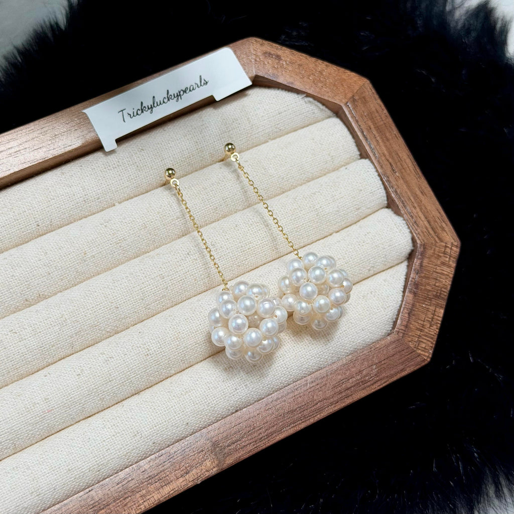 A pair of gold and white pearl dangle earrings displayed on a cream fabric-lined wooden jewelry box. The earrings feature small white pearls clustered in round flower-like shapes, hanging from delicate gold chains.