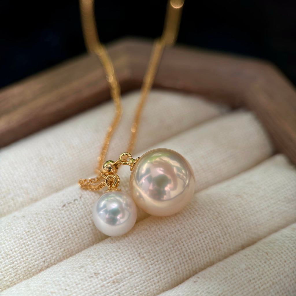 A gold necklace with two pearl-like pendants, one larger and one smaller, displayed on a cream fabric jewelry stand.