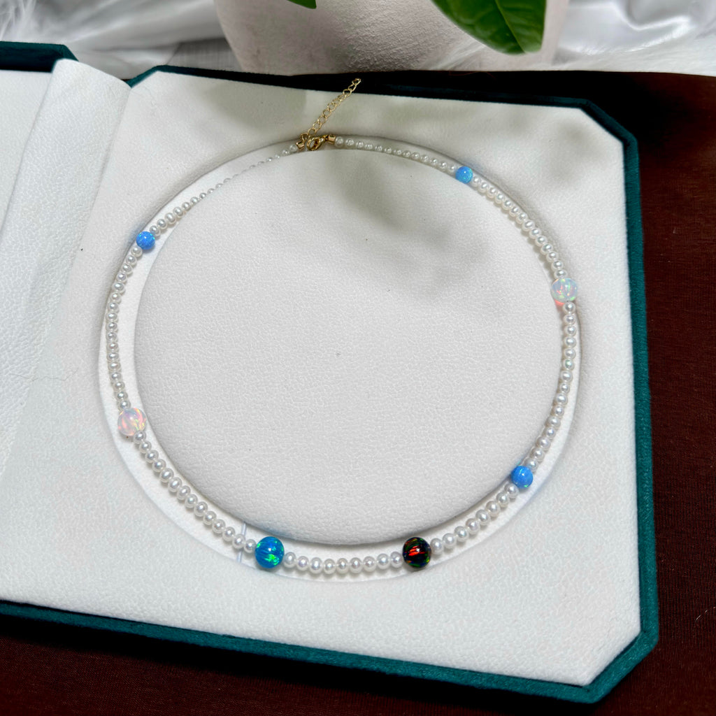 A delicate double-strand pearl necklace with small blue and iridescent beads, displayed inside an open white jewelry box with a dark green velvet border.