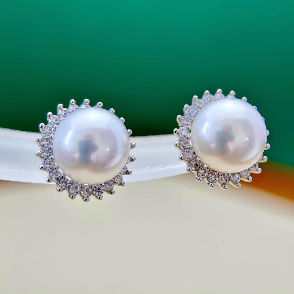 A pair of round pearl stud earrings with silver-toned halo settings, displayed on a white curved stand against a green and cream background.