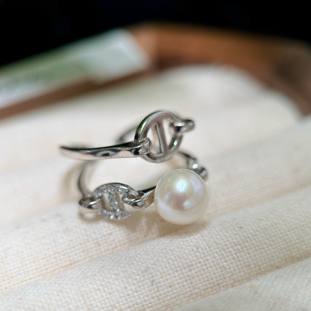 A silver-toned ring with a round white pearl and a small clear crystal detail, displayed on a textured cream fabric surface.