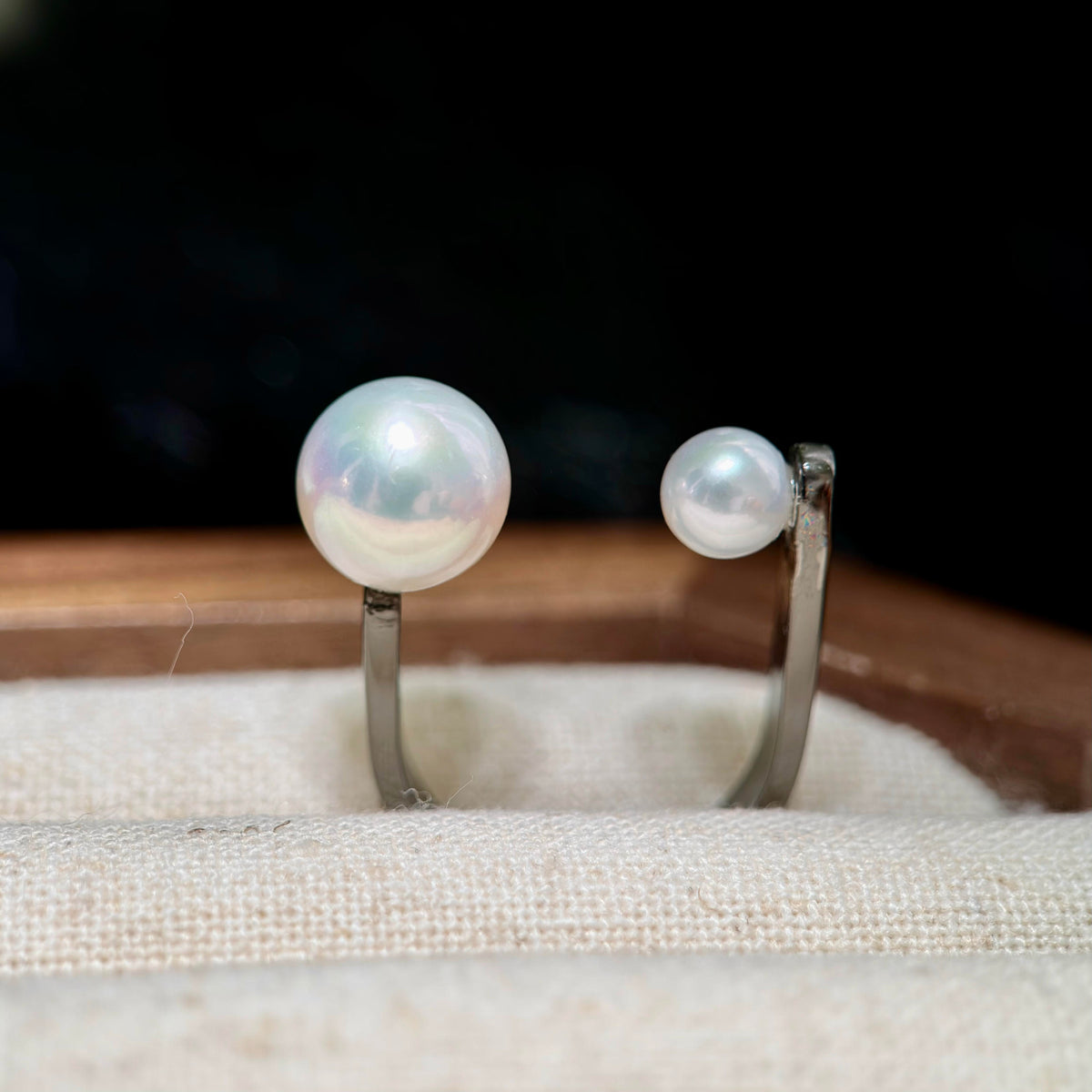 A pair of silver ring posts with large and small round white pearls, displayed on a textured beige fabric surface with a dark background.