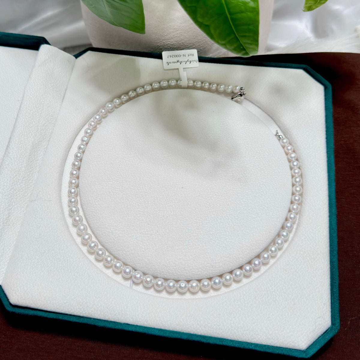 A single-row pearl necklace with silver-toned metal findings, displayed inside an open white jewelry box with a dark green velvet border.