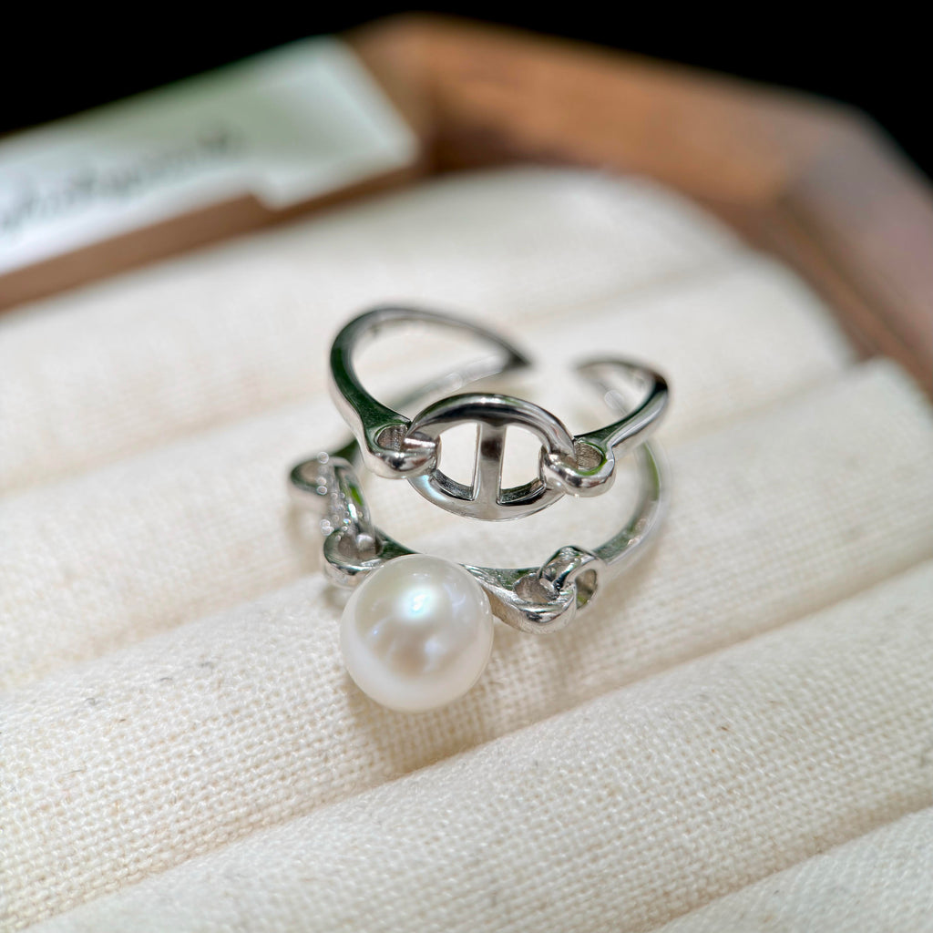 A silver-toned open ring with a single round white pearl hanging from it, displayed on a cream fabric jewelry cushion.
