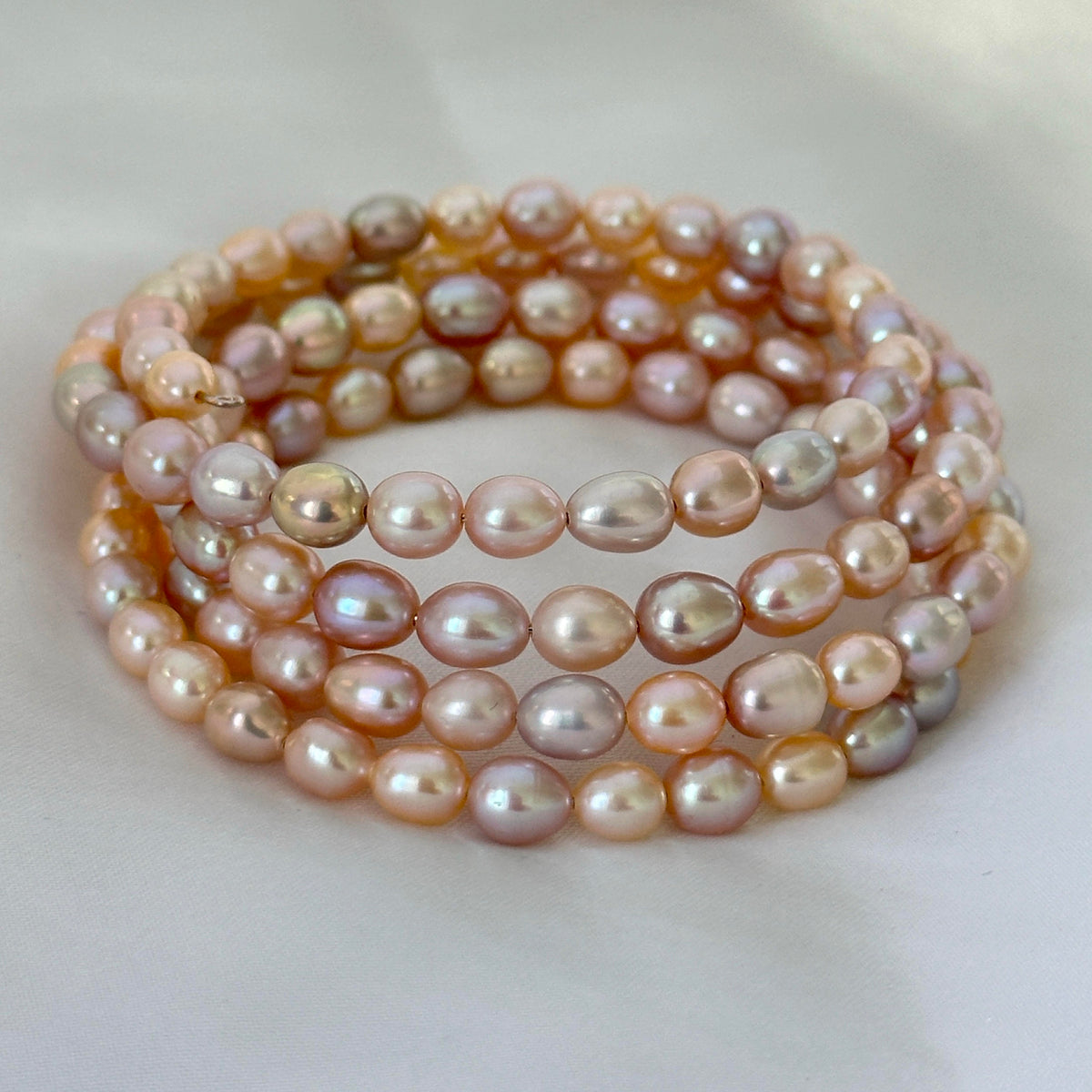 A stack of five beaded stretch bracelets made of iridescent freshwater pearls in soft peach, pink, and cream tones, arranged on a light off-white background.