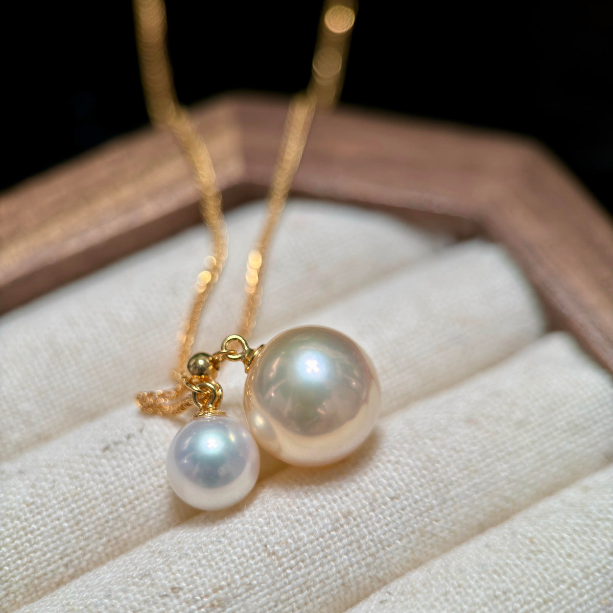 A gold chain necklace with two pearl-like drops, one larger pearlescent white pearl and one smaller white pearl, displayed on a cream fabric jewelry cushion.