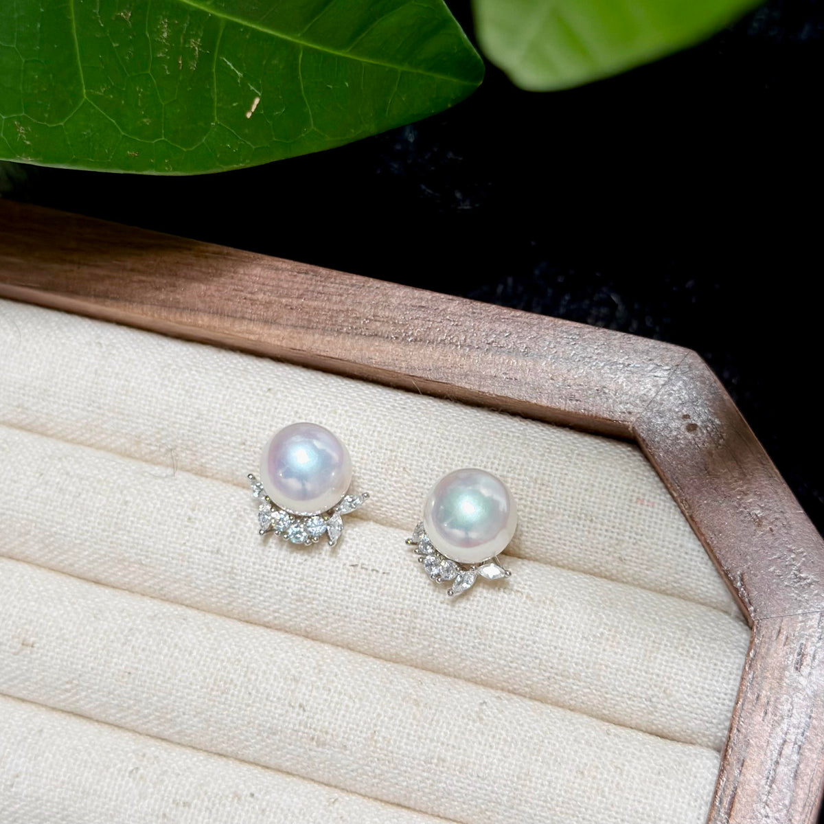 A pair of pearl stud earrings with small clear crystal leaf accents, displayed on a cream fabric-lined jewelry box with a wooden border, with green leaves in the background.