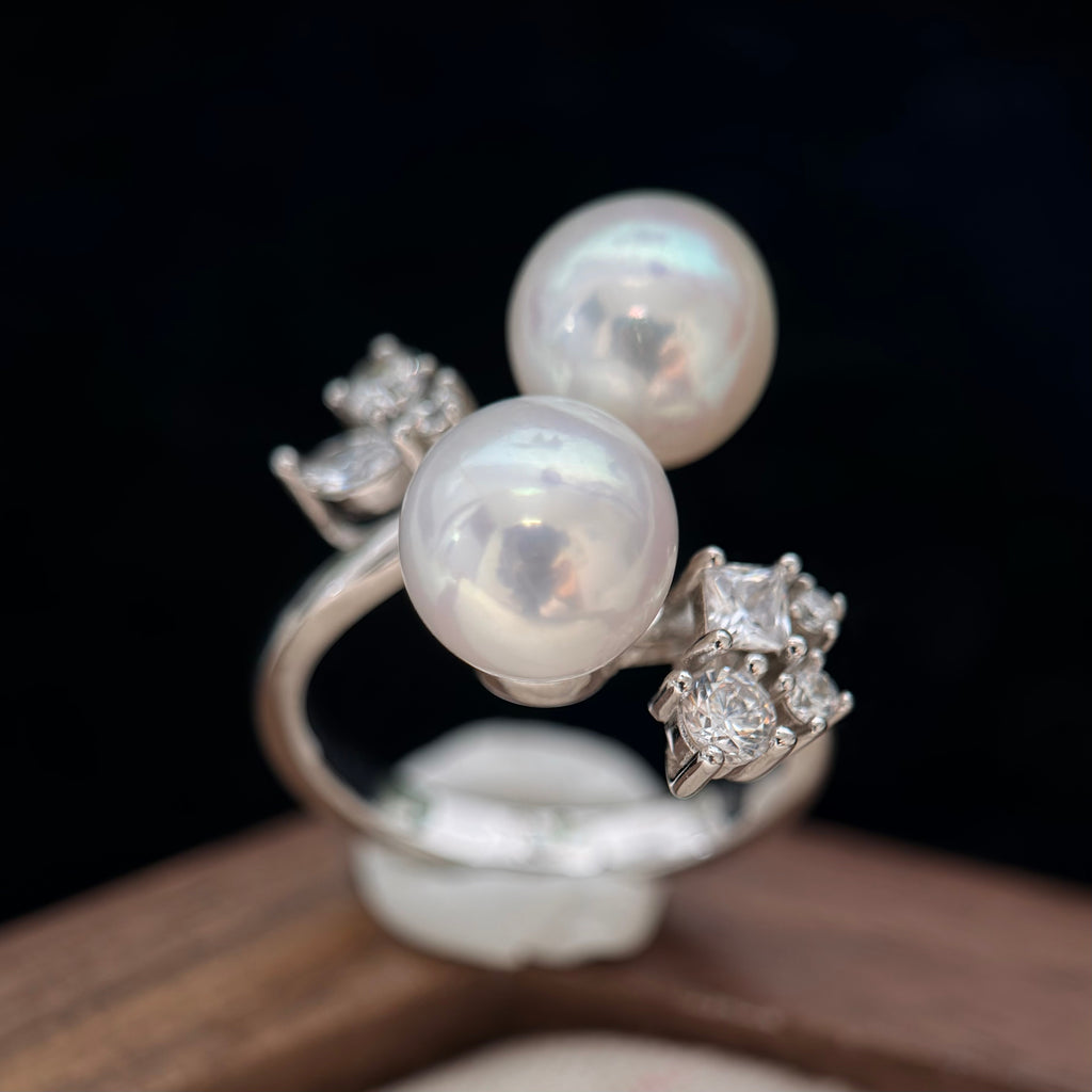 A silver-toned ring featuring two large round white pearls and small clear gemstone clusters, photographed close-up against a dark background.