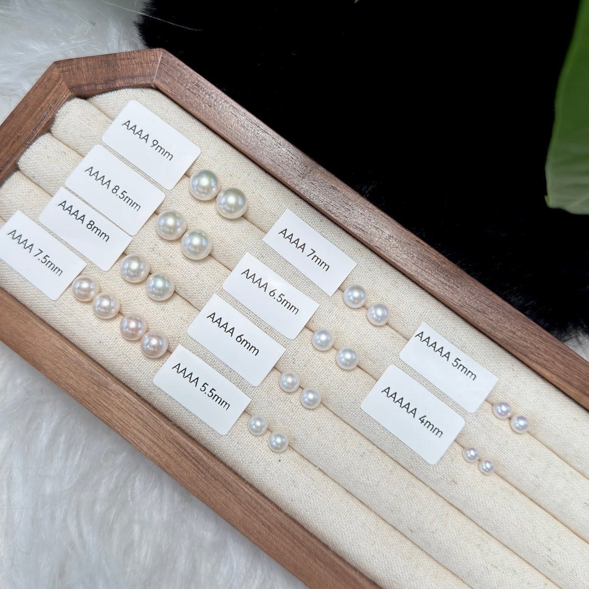 A wooden display tray with cream fabric rolls holding small pearl bead strands labeled by size in millimeters, arranged on a white furry surface.