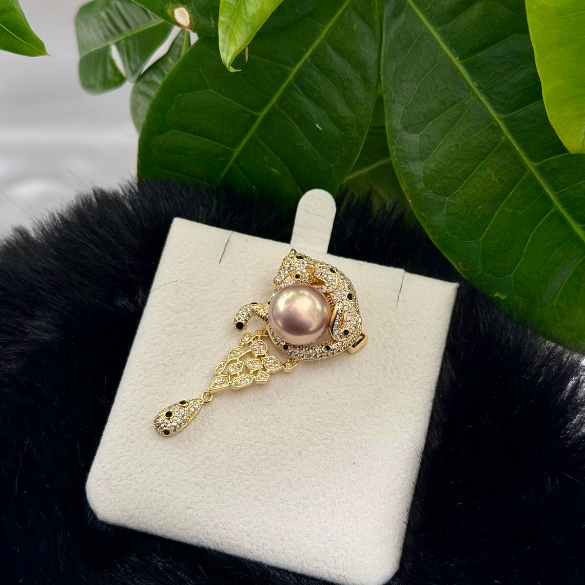 A gold-toned ornate brooch featuring a central pink pearl and small clear stones, displayed on a white jewelry card over black faux fur with green leaves in the background.