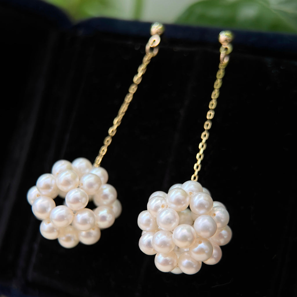A pair of gold dangle earrings with round white pearl clusters at the bottom, displayed on a black velvet jewelry box.