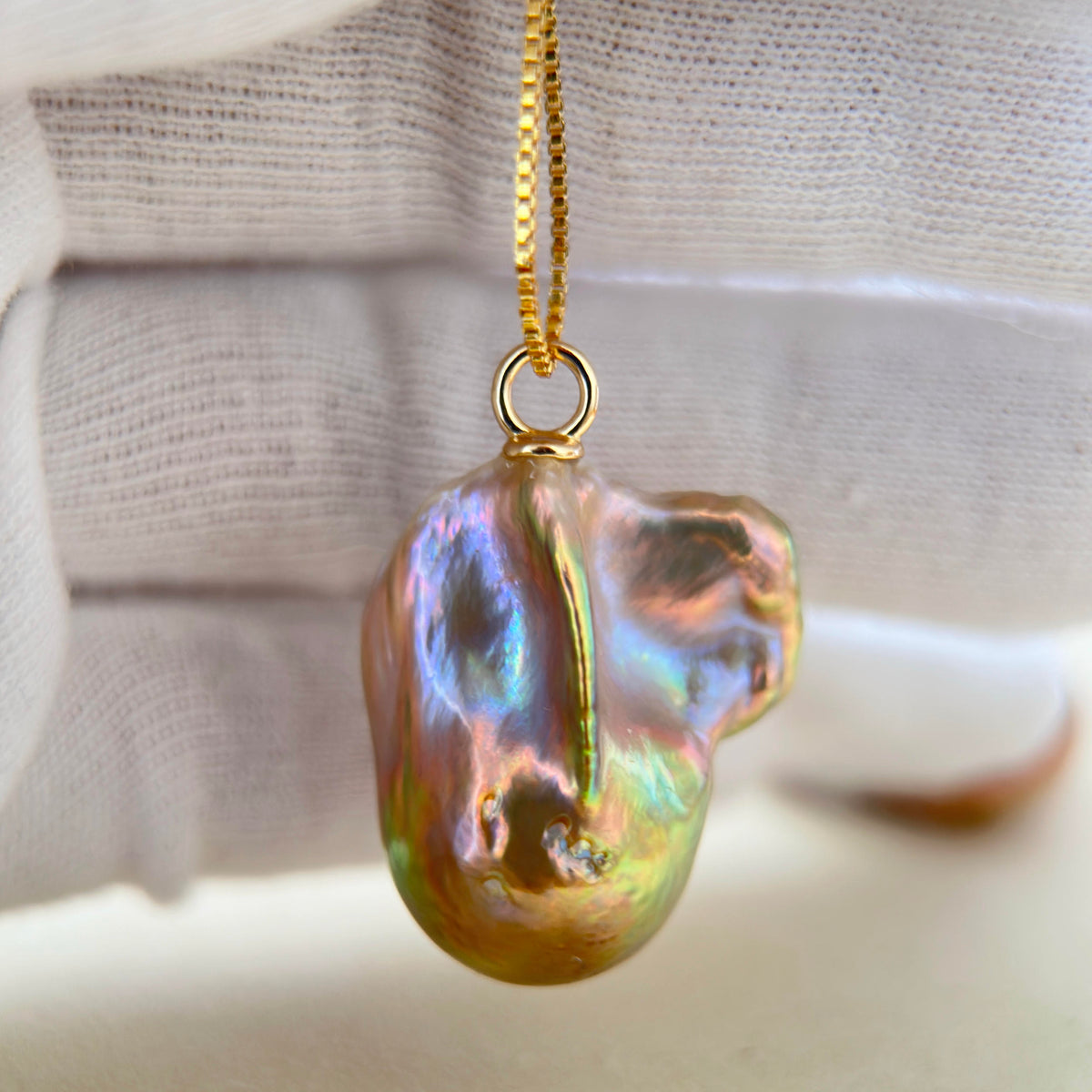 A close-up of a gold chain necklace with a baroque pearl pendant. The pearl has a pearlescent, iridescent finish reflecting shades of green, blue, and gold, suspended from a gold ring on a delicate gold chain, set against a soft, textured white fabric background.