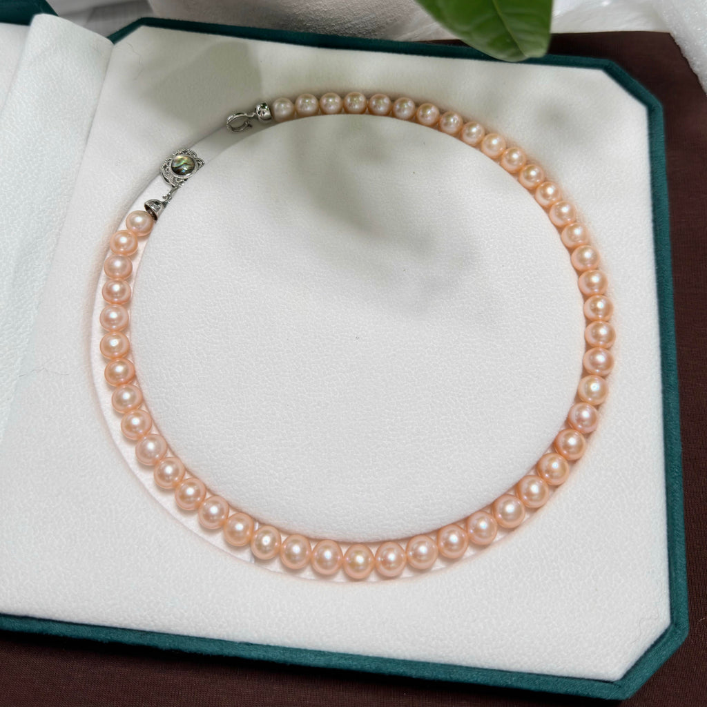 A strand of light golden freshwater pearls arranged in a circle on a white jewelry display pad with a dark green border. The necklace has a silver-toned clasp with a small green stone accent.
