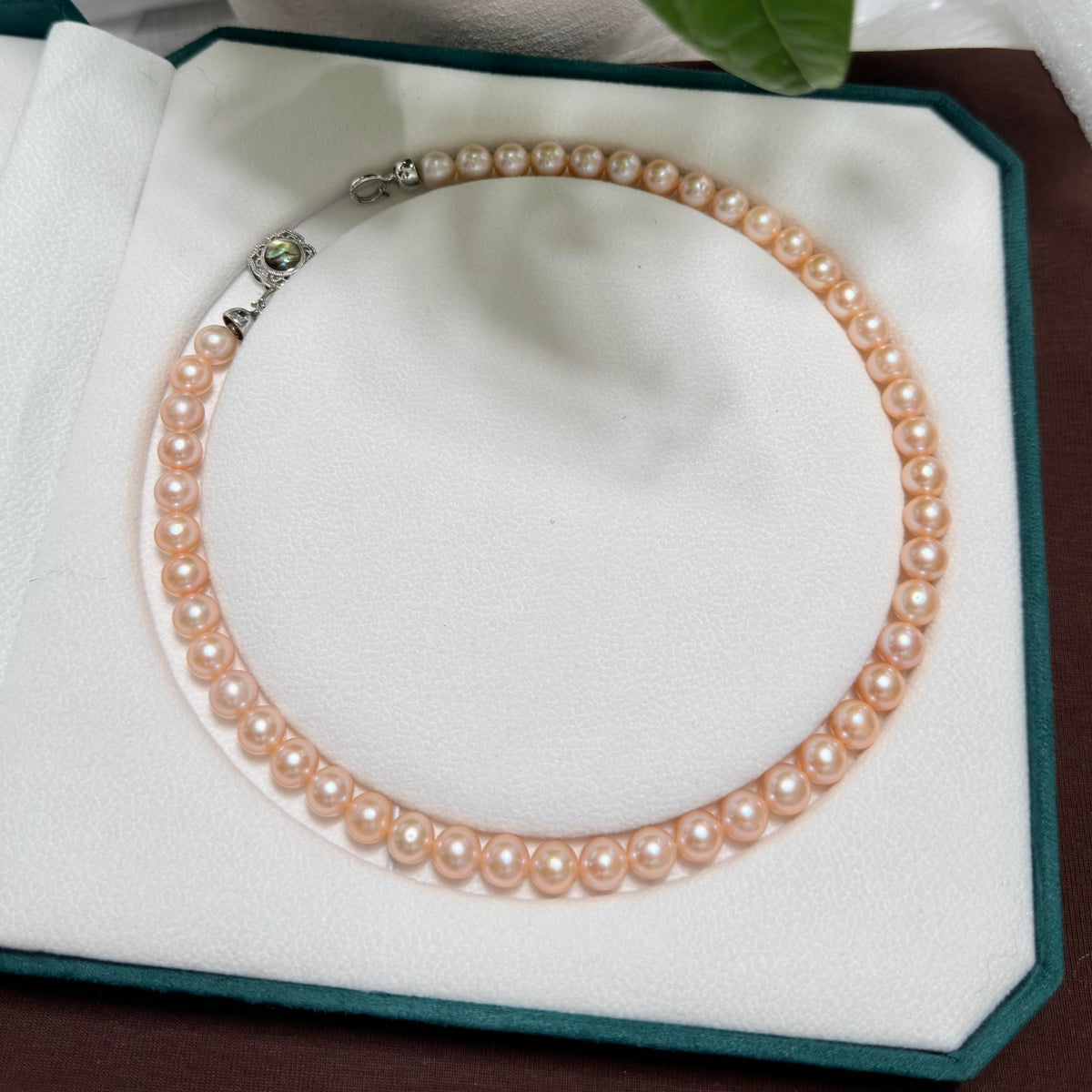 A strand of light golden freshwater pearls arranged in a circle on a white jewelry display pad with a dark green border. The necklace has a silver-toned clasp with a small green stone accent.