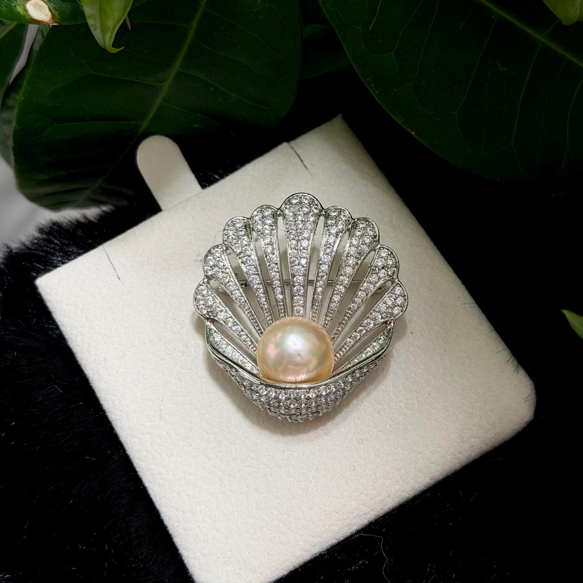 A shell-shaped silver brooch with a central iridescent pearl and pavé-set clear stones, displayed on a white jewelry stand with dark green leaves in the background.