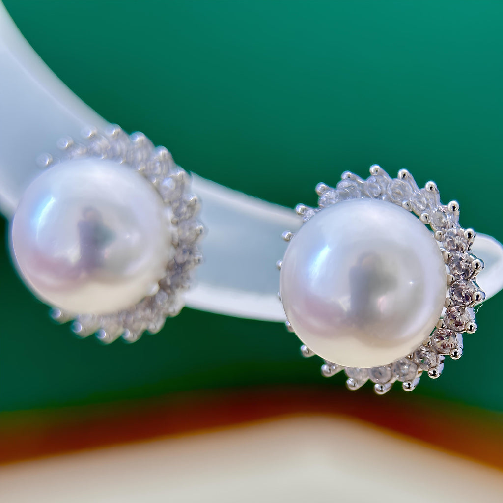 A close-up of a pearl stud earring with a silver-toned halo of small clear stones, mounted on a translucent display stand against a green background.