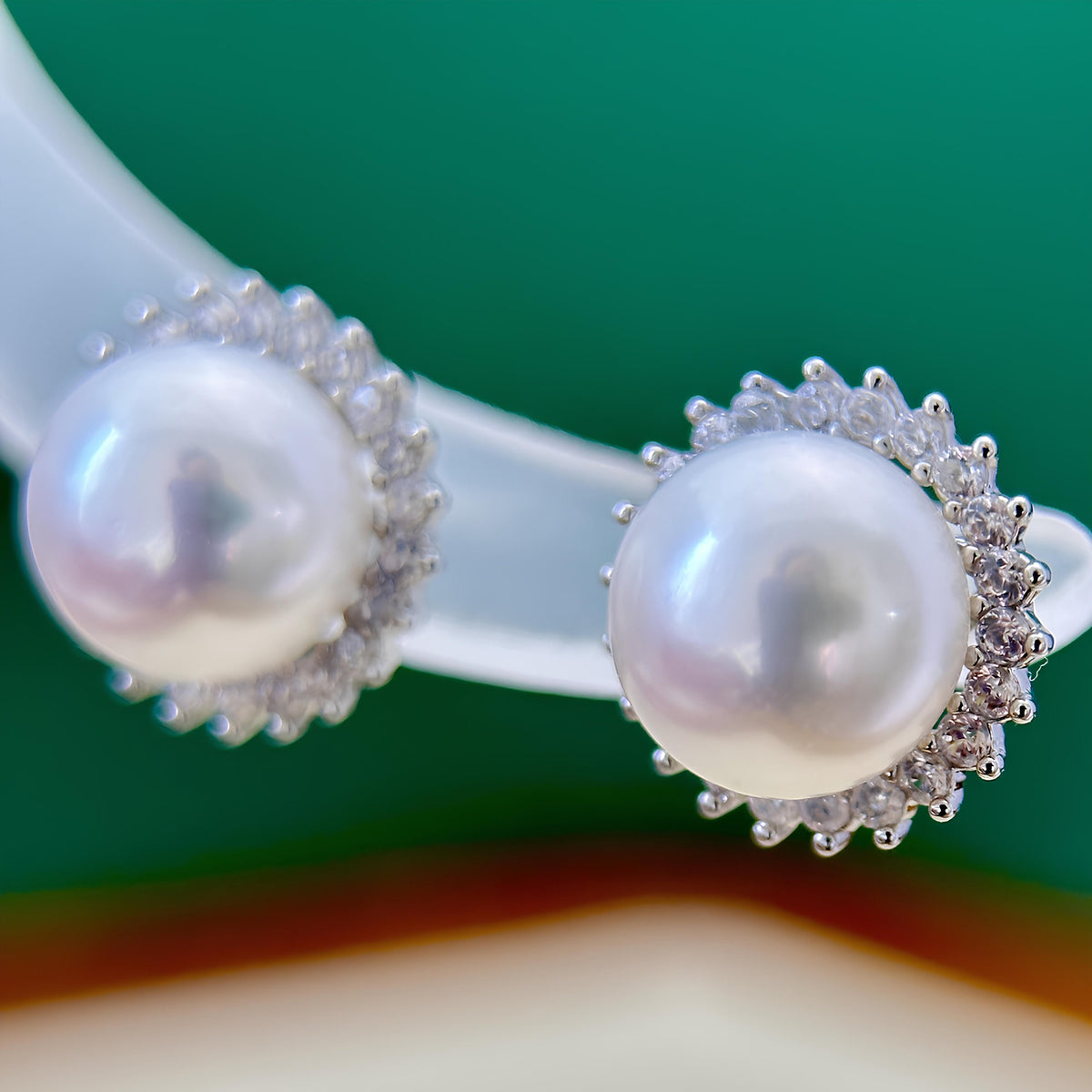 A close-up of a pearl stud earring with a silver-toned halo of small clear stones, mounted on a translucent display stand against a green background.