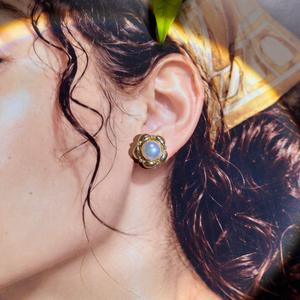 A close-up of a person's ear wearing a gold flower-shaped stud earring with a round white pearl center, set against dark hair and a patterned headscarf.