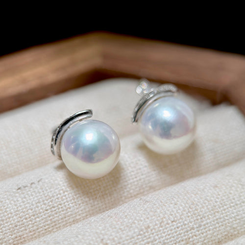 A pair of silver stud earrings with round white freshwater pearls, displayed on a textured cream fabric surface with a wooden tray edge in the background.