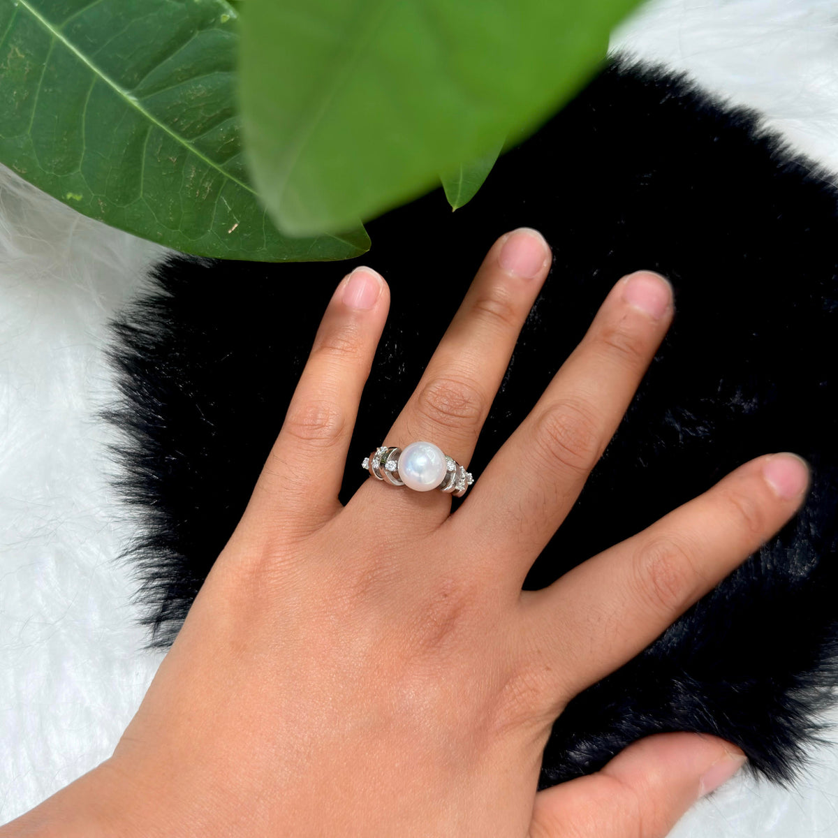 A silver ring with a large white pearl and small clear stones on the ring finger of a hand, resting on a black furry surface with a green leaf in the background.