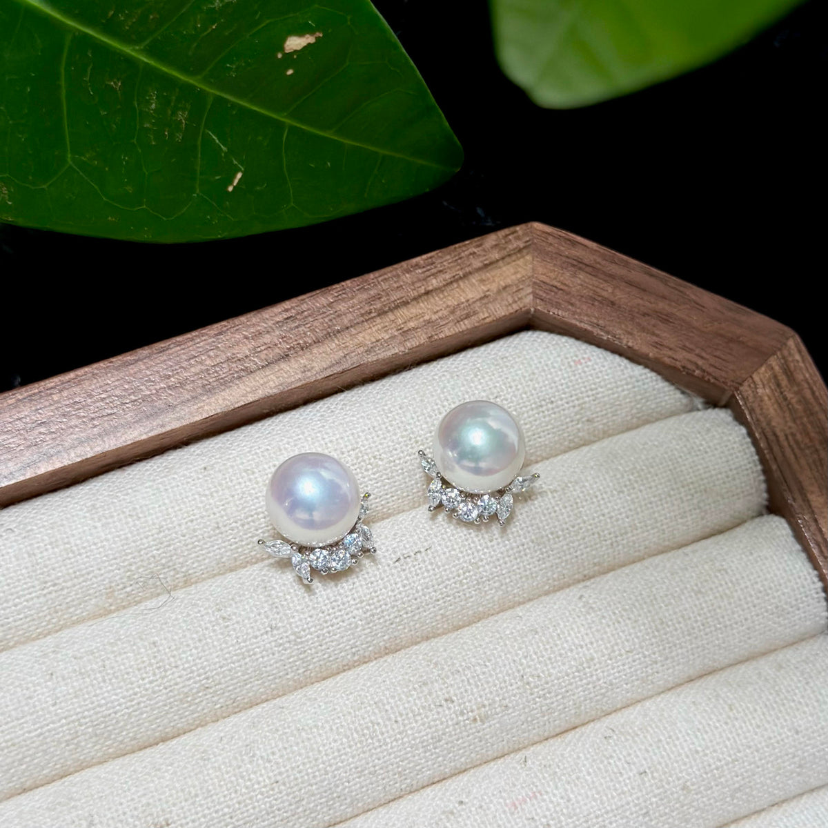 A pair of pearl stud earrings with silver floral settings and small clear stones, displayed on a cream fabric-lined jewelry box with a wooden edge. Green leaves are visible in the background against a black backdrop.