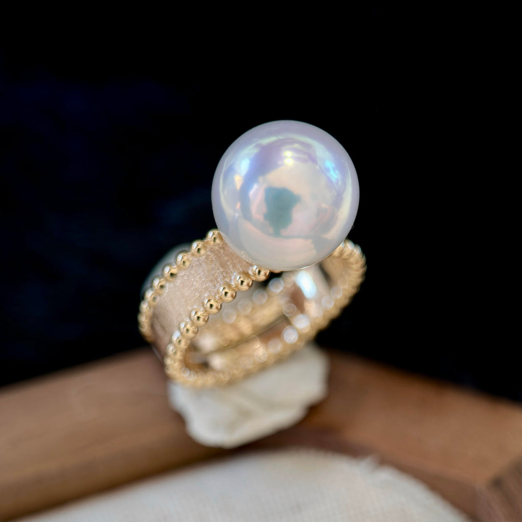A gold ring featuring a large, lustrous white pearl on top, displayed on a small stand against a dark background.