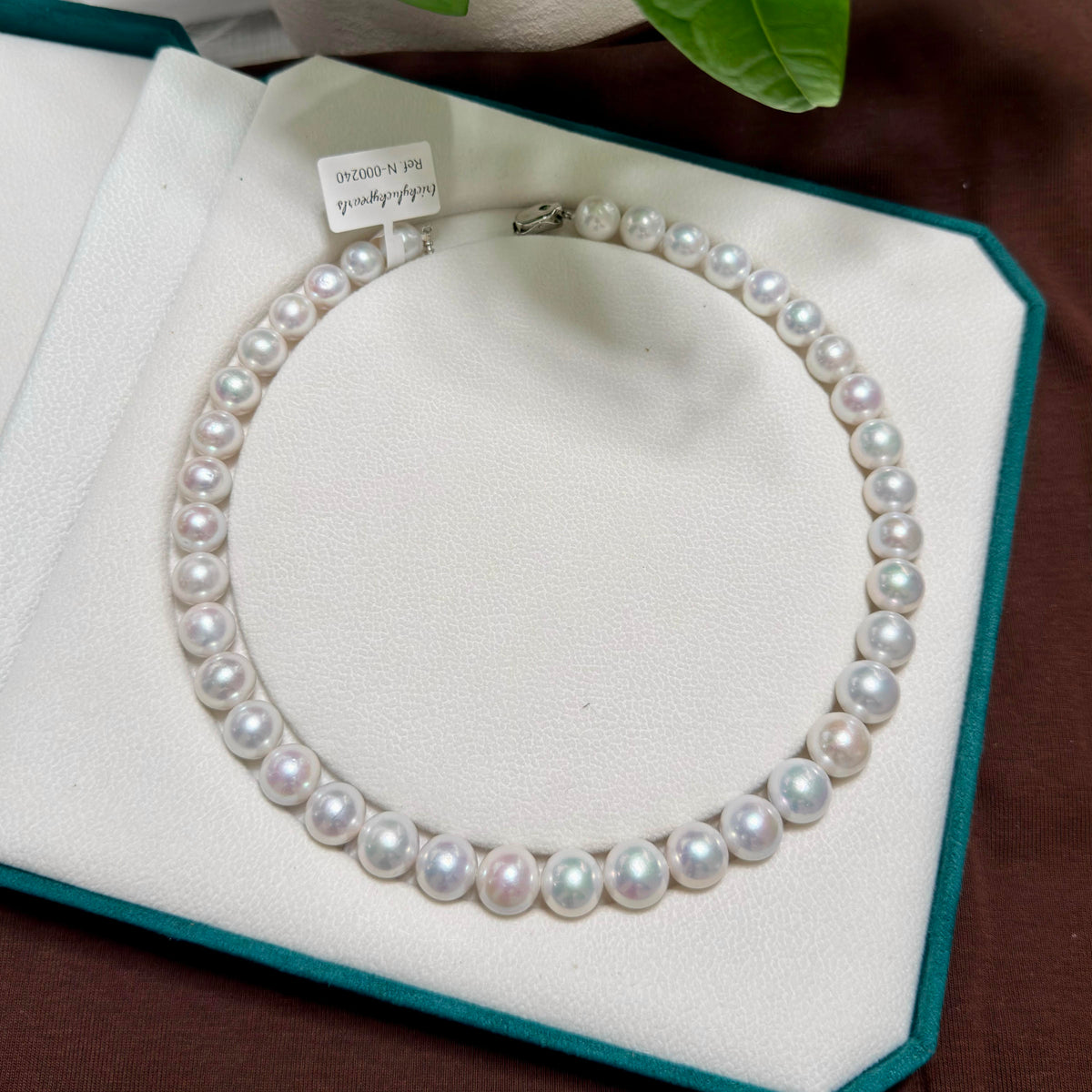 A strand pearl necklace arranged in a circle on a white jewelry display cushion inside an open teal box. The pearls are white with soft pearly luster and are secured by a silver clasp.
