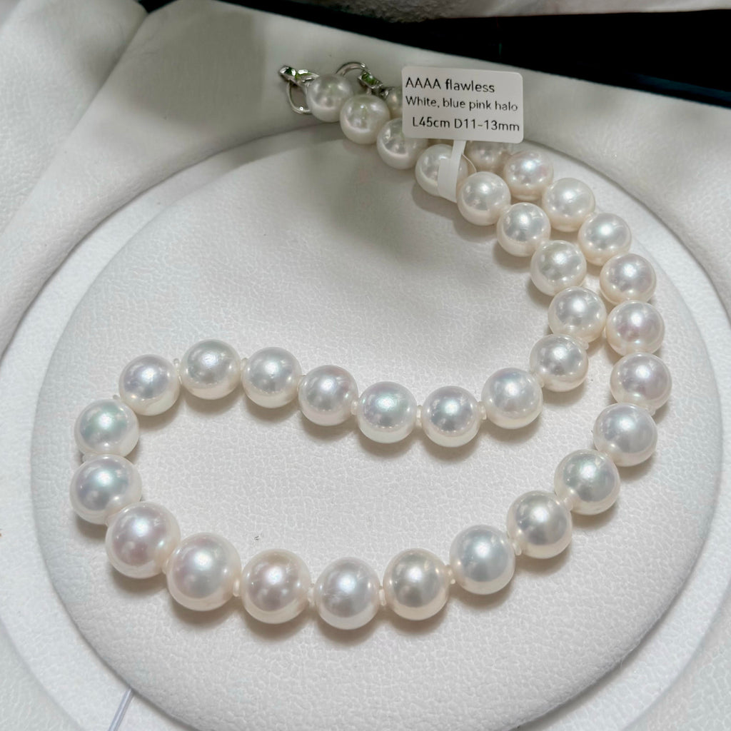 A strand of white blue pink pearl beads arranged in a circle on a white circular display cushion, with a small paper tag attached near the clasp.