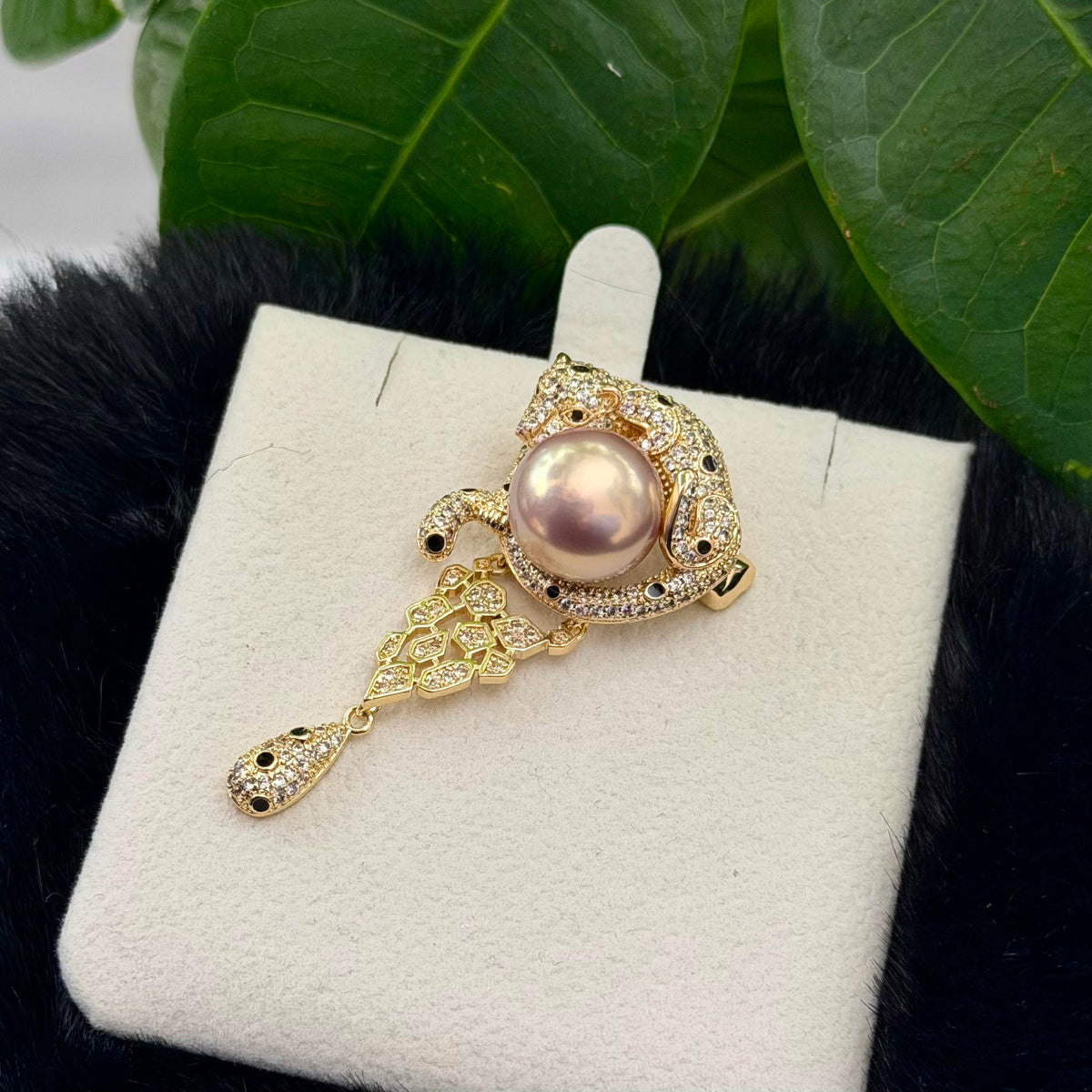 A gold-toned pendant brooch featuring a central pinkish pearl and pavé-set stones, displayed on a white jewelry stand with dark green leaves and black faux fur in the background.