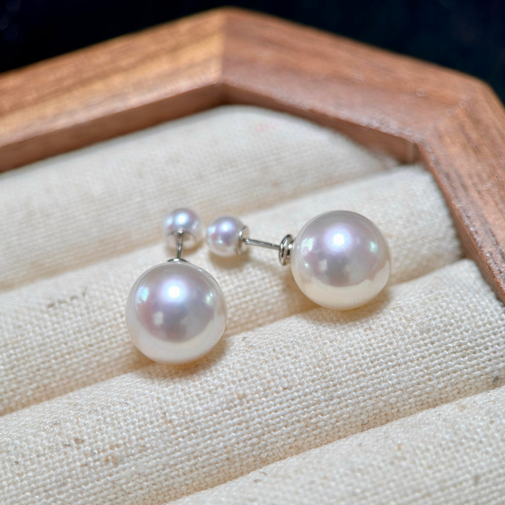 A pair of pearl stud earrings with small matching pearl studs, displayed on a cream fabric jewelry cushion inside a wooden box.