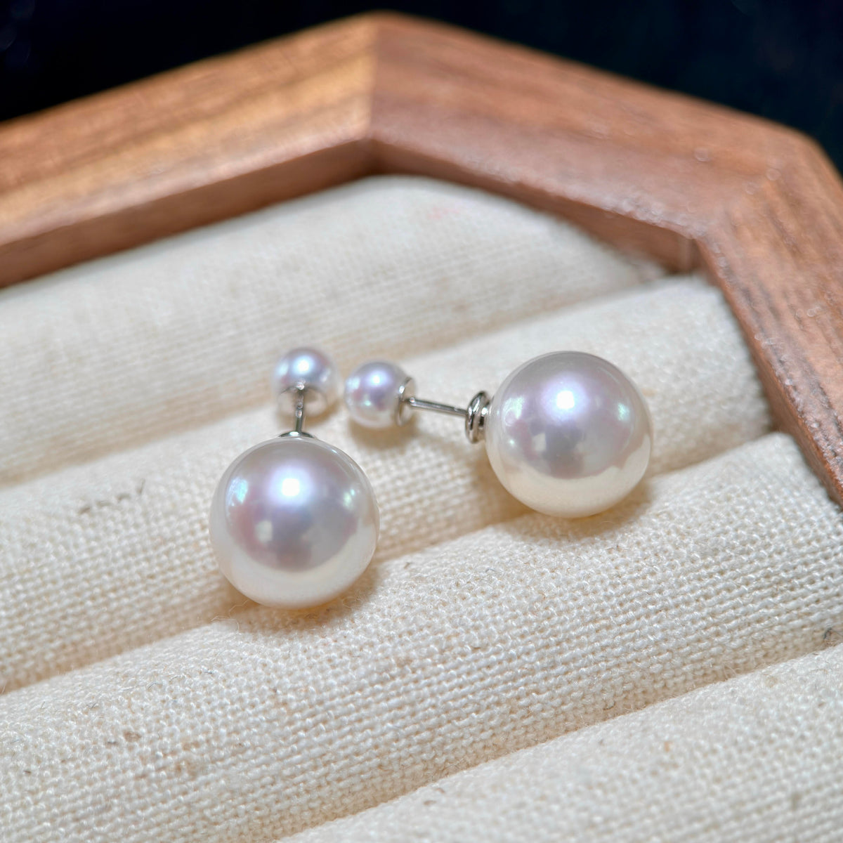 A pair of pearl stud earrings with small matching pearl studs, displayed on a cream fabric jewelry cushion inside a wooden box.
