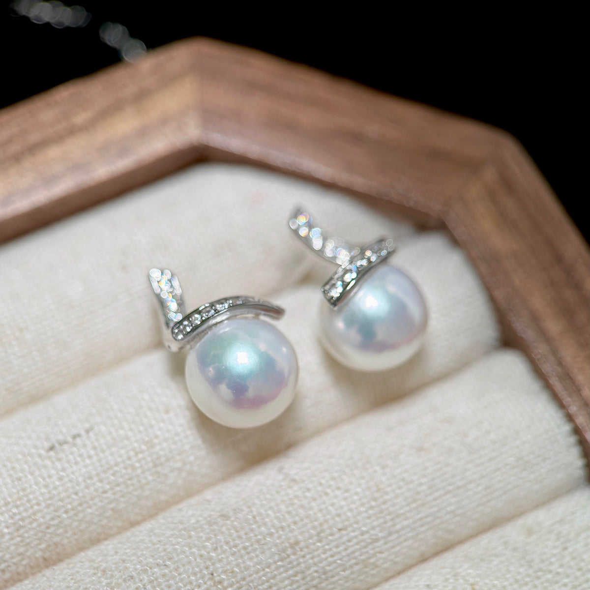 A pair of silver stud earrings with round white freshwater pearls and small clear stones, displayed on a cream fabric-lined jewelry box inside a wooden frame.