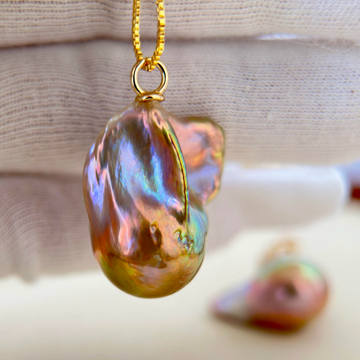 A close-up of a gold pendant necklace featuring a large, baroque-shaped iridescent shell bead that shifts between pink, green, and gold tones, suspended from a gold chain against a soft beige fabric background.