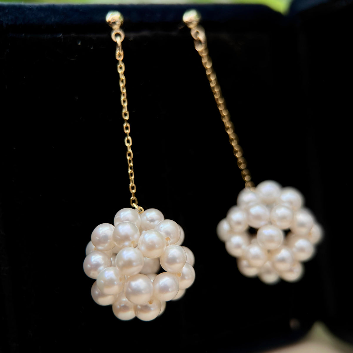 A pair of gold drop earrings with spherical clusters of white freshwater pearls, displayed on a black velvet jewelry box.