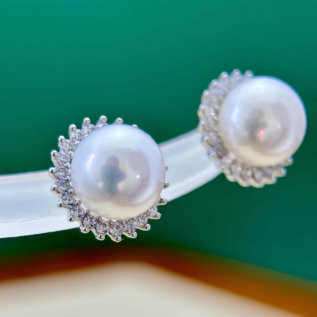 A pair of pearl stud earrings with silver-toned sunburst settings and small clear stones, displayed on a clear stand against a green background.