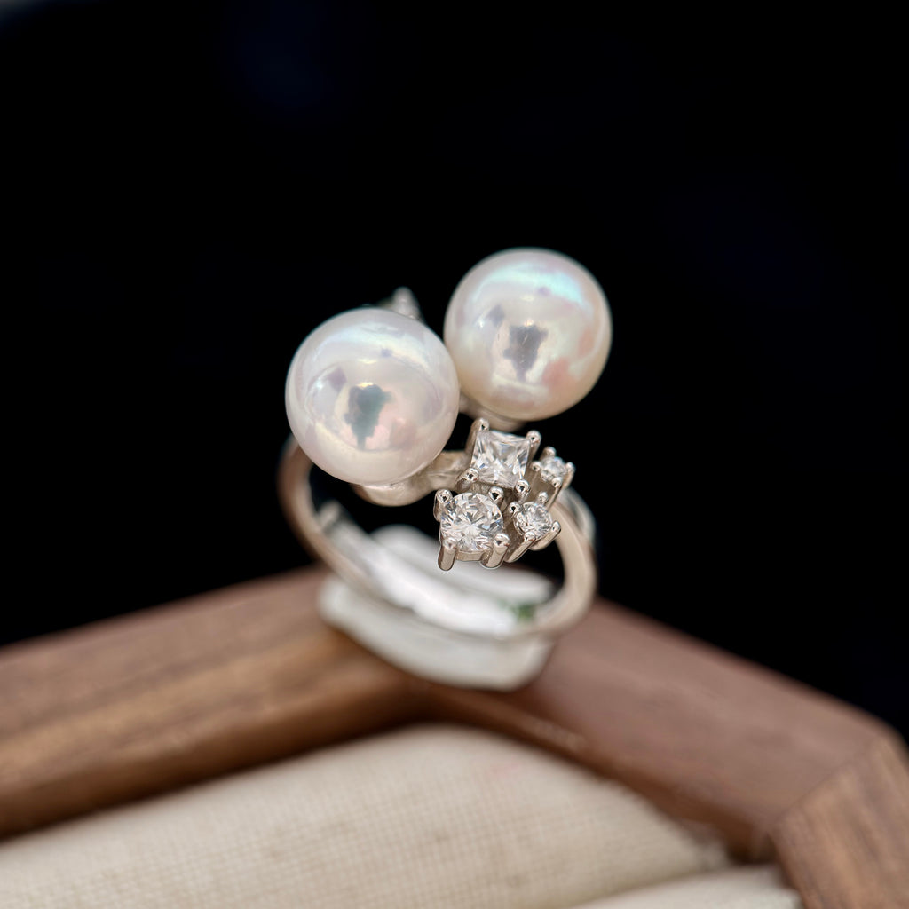 A silver ring featuring two large white pearls and a small cluster of clear stones, displayed on a wooden surface with a dark background.