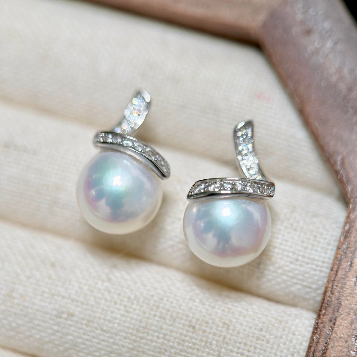 A pair of pearl stud earrings with silver-toned settings and small clear stones, displayed on a cream textured jewelry box.