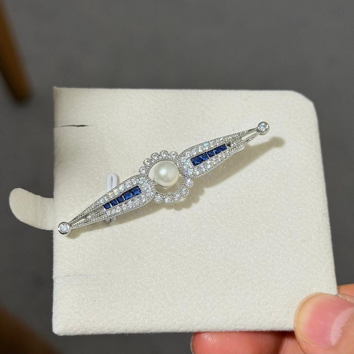 An Art Deco style silver-toned brooch featuring a central white pearl, surrounded by clear pavé stones and accented with blue baguette-cut stones, displayed on a cream jewelry card held by a hand.