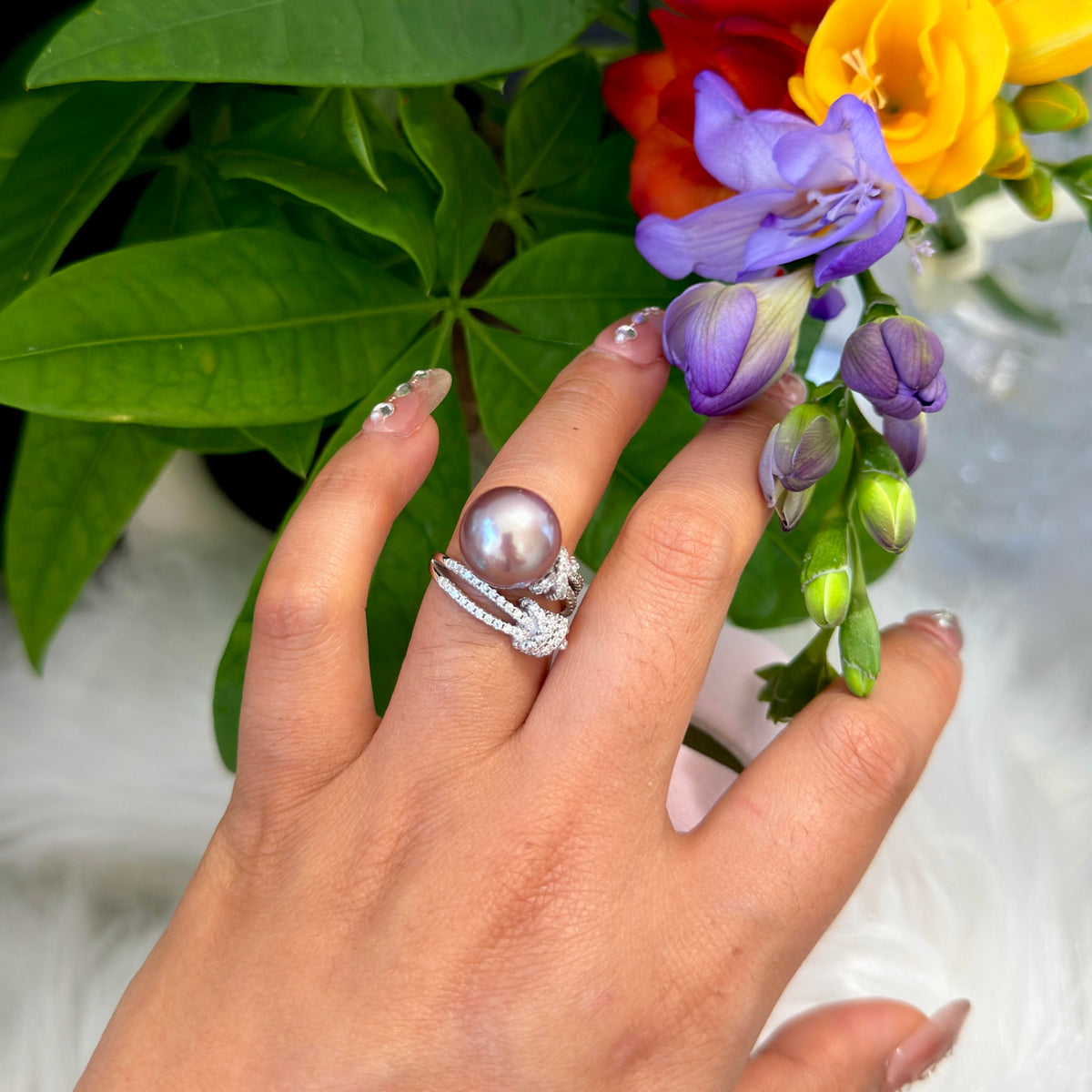 A close-up of a hand wearing a silver ring with a large round purple blue pearl and a smaller diamond-accented band, posed against a background of green leaves and colorful flowers.