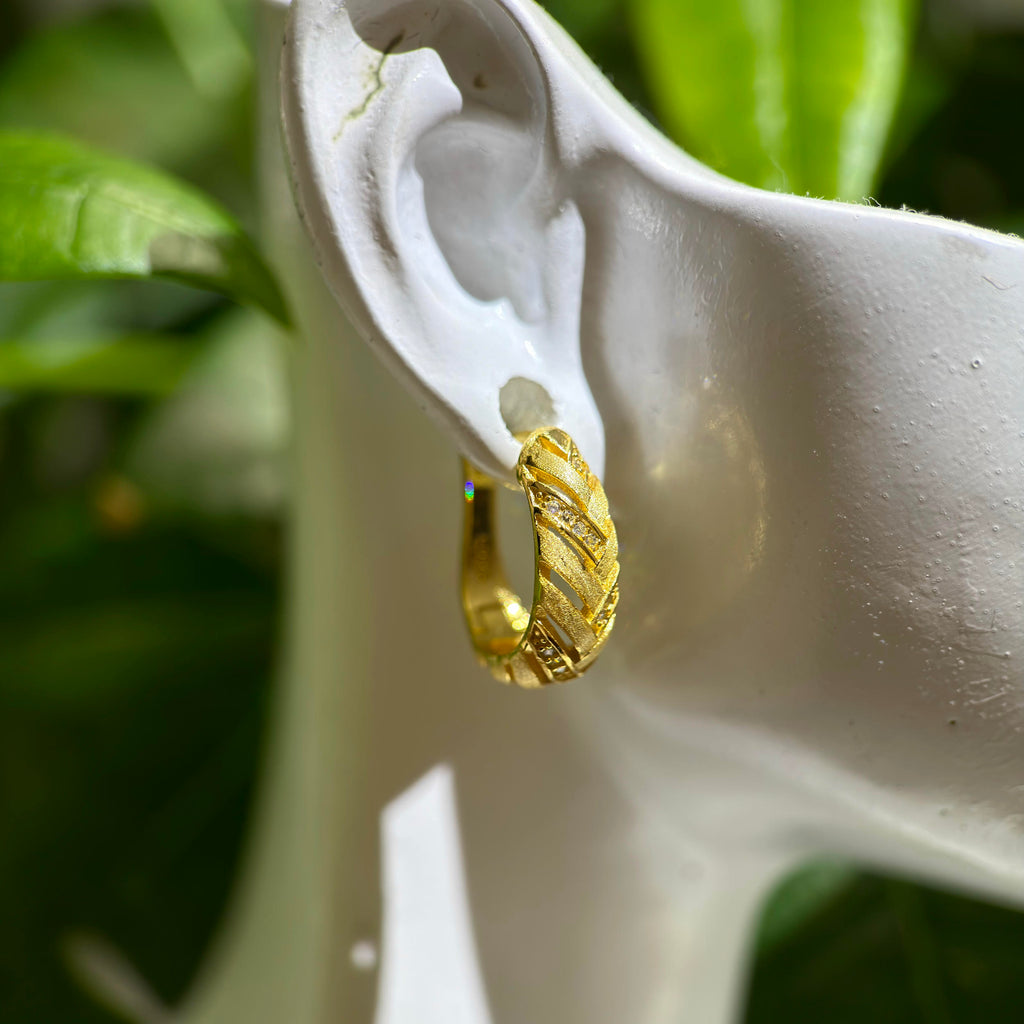 A gold huggie hoop earring with a textured, ribbed pattern, shown on a white ear display form against a background of green leaves.