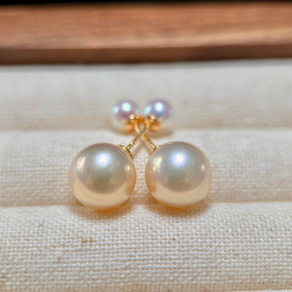 A pair of gold stud earrings with large, round, lustrous pearl-like beads in a warm cream or ivory tone, displayed on a textured beige fabric surface.