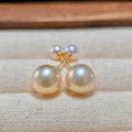 A pair of gold stud earrings with large, round, lustrous pearl-like beads in a warm cream or ivory tone, displayed on a textured beige fabric surface.