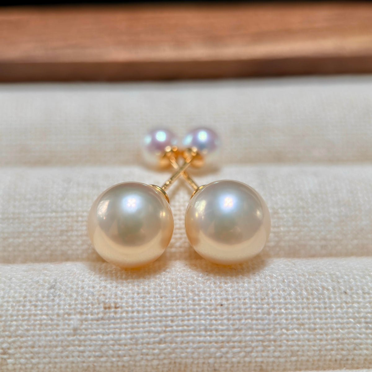 A pair of gold stud earrings with large, round, lustrous pearl-like beads in a warm cream or ivory tone, displayed on a textured beige fabric surface.