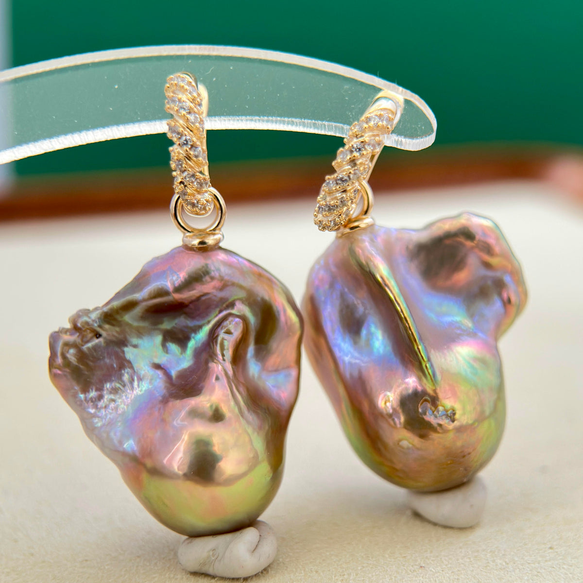 A pair of gold baroque pearl earrings with twisted gold wire hooks and small clear stones, displayed on a clear stand against a green and beige background.