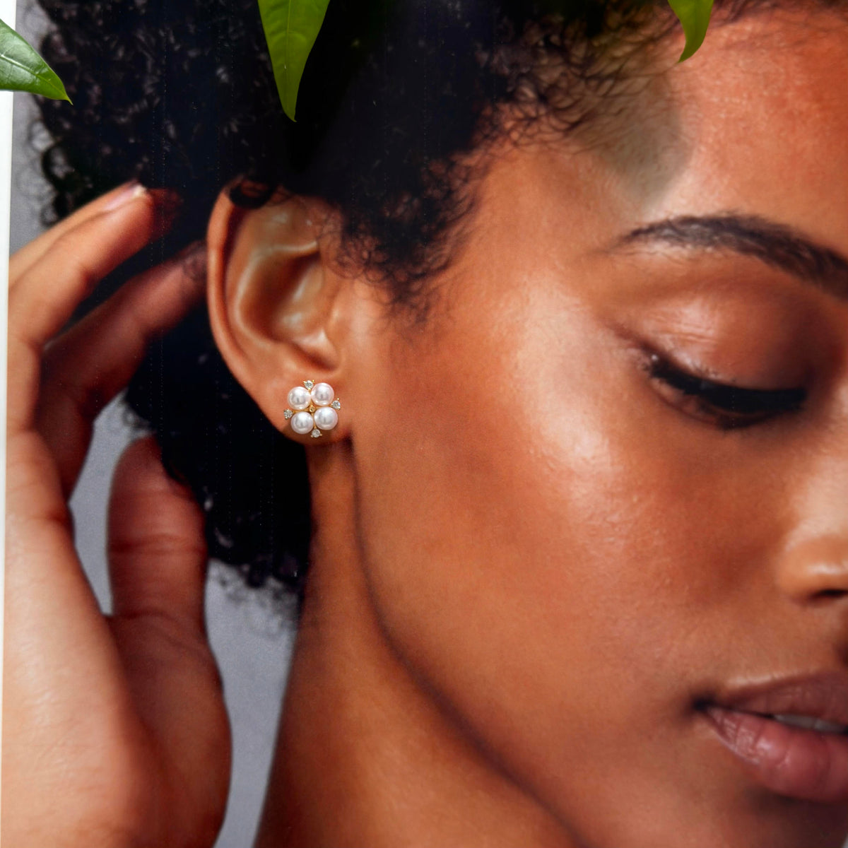 A close-up of a woman wearing a small flower-shaped stud earring made of white pearls with a silver center, shown against her ear with her hand near her hair.