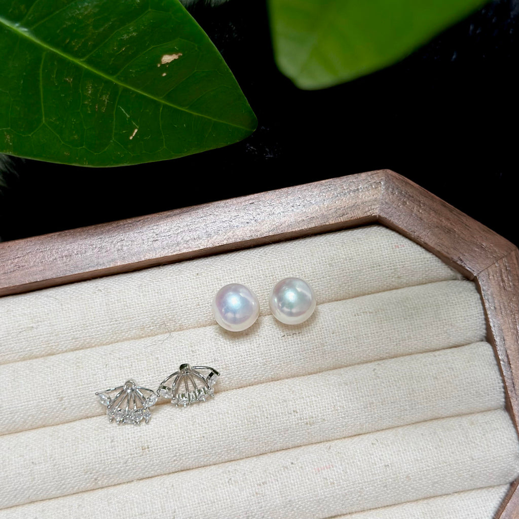 A pair of white freshwater pearl stud earrings and a pair of silver shell-shaped stud earrings displayed on a cream fabric-lined jewelry box, with green leaves in the background.
